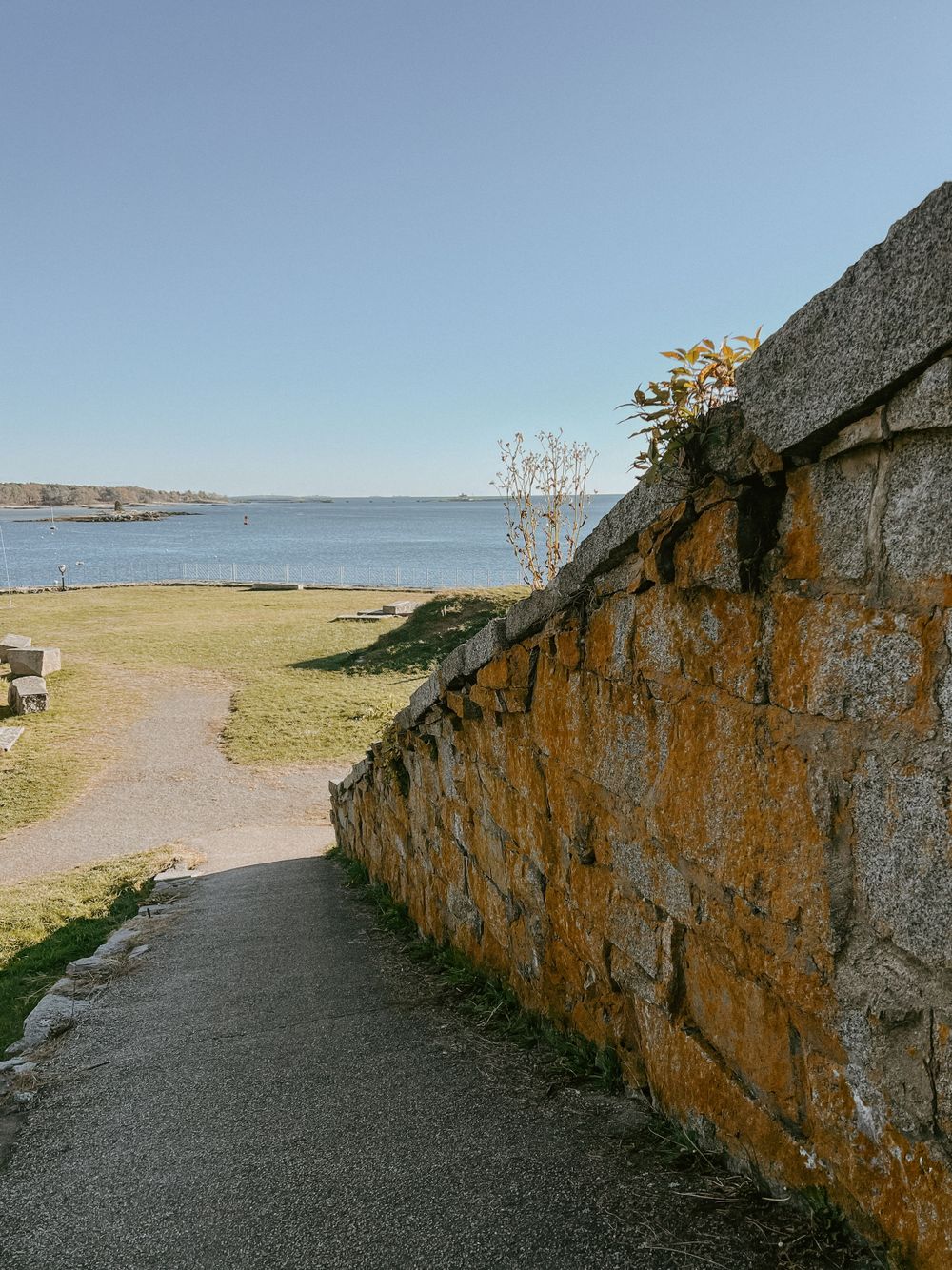 A curved stone wall path leads to a waterfront view on a sunny day.
