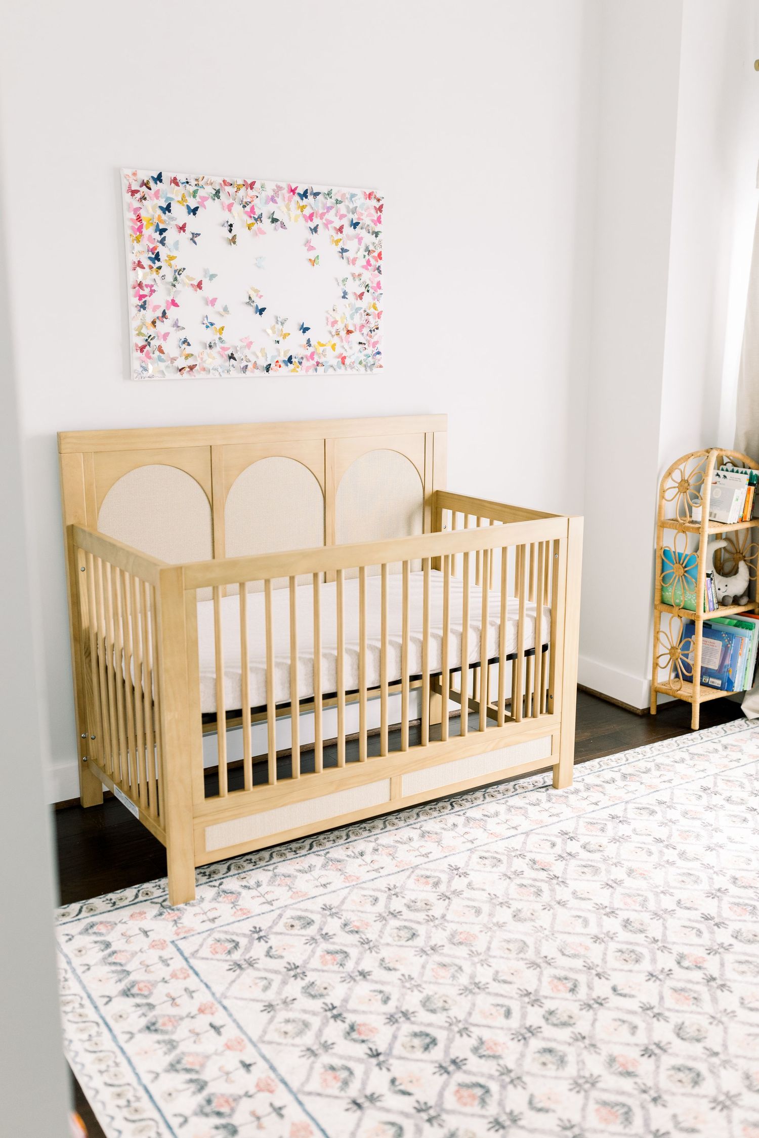 Natural wood crib with butterfly artwork in a bright nursery with patterned rug.