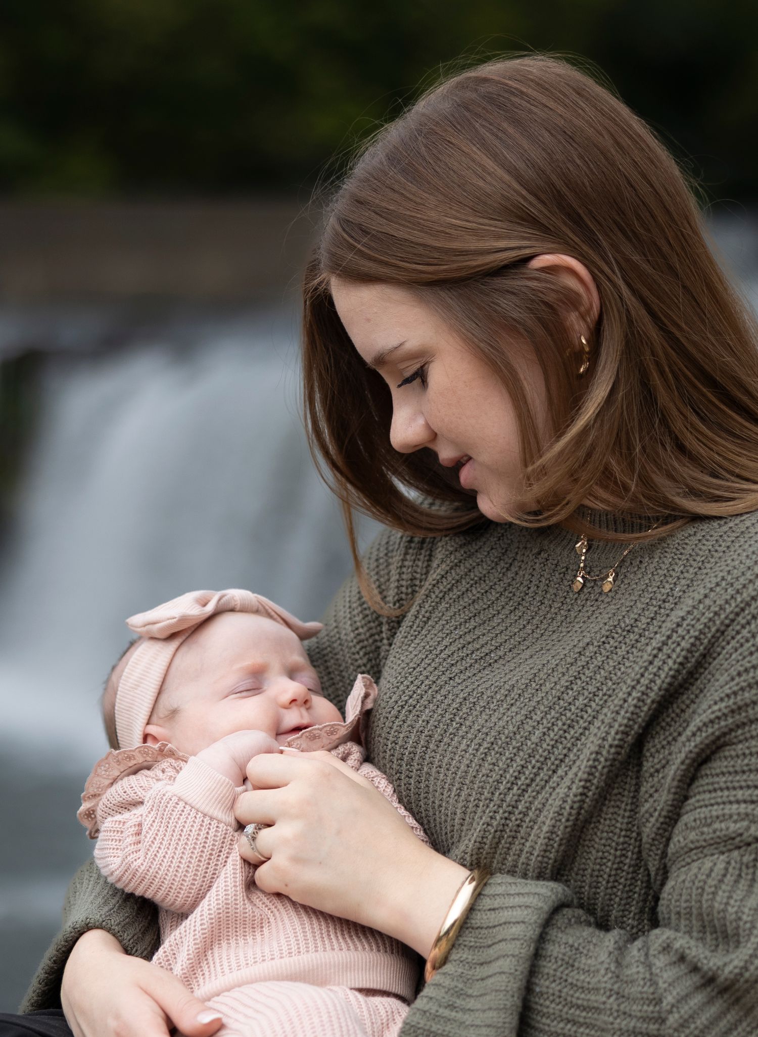 Tender moment between parent and sleeping baby in pink against waterfall background.