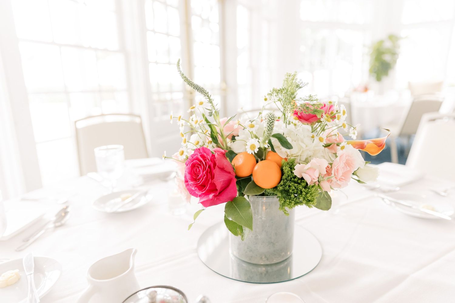 Fresh floral centerpiece with pink roses and citrus in a metal container on an elegant white table setting.