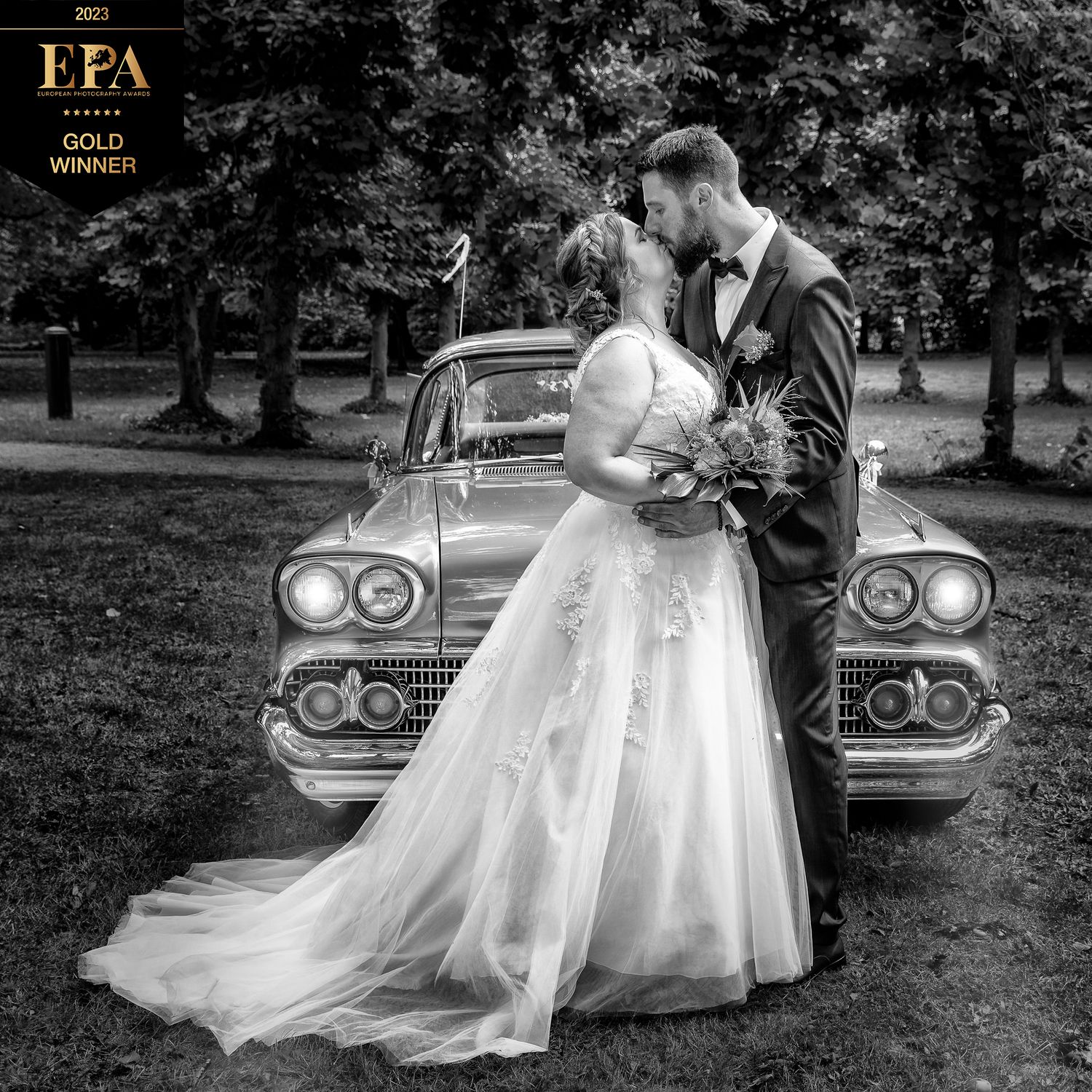 Newlywed couple poses romantically by vintage car in black and white wedding photograph.