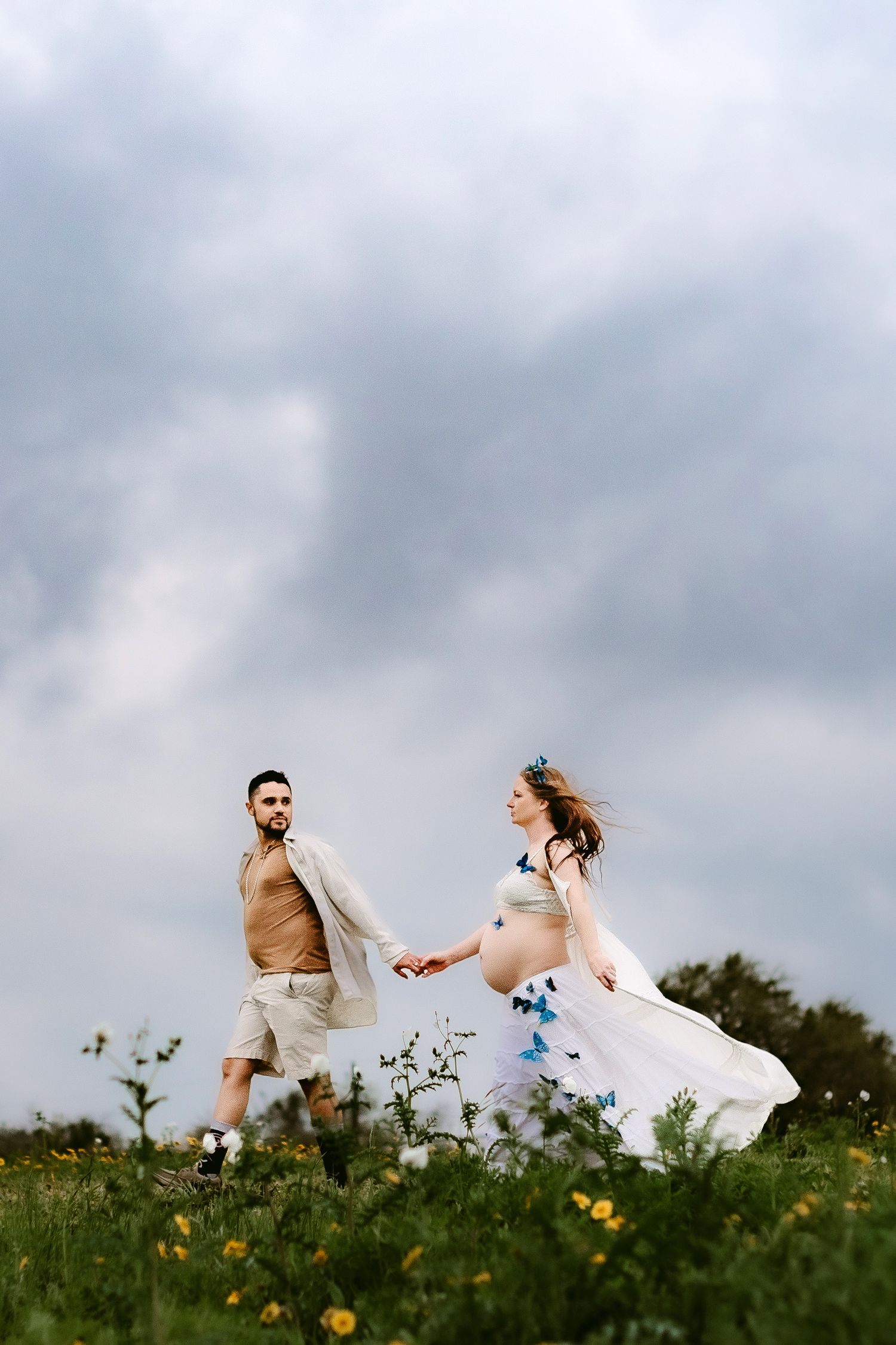 A couple runs through a wildflower meadow under dramatic cloudy skies during a romantic outdoor photoshoot.