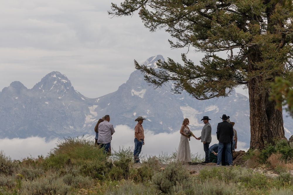 Outdoor wedding ceremony with couple holding hands near a pine tree, misty Grand Teton mountains in the background.
