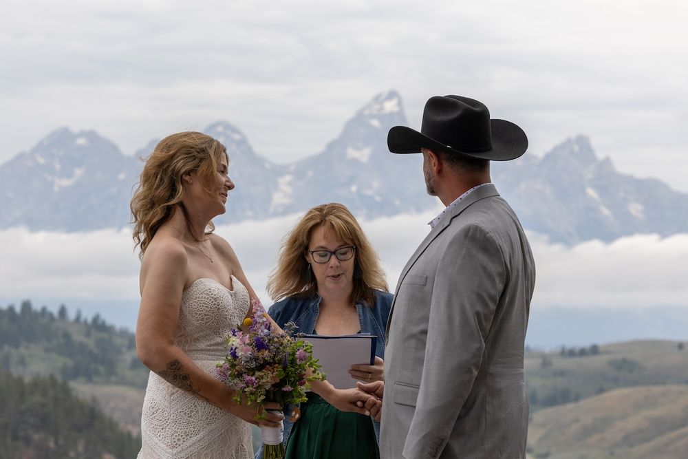Outdoor wedding ceremony with couple exchanging vows before Grand Teton mountains, officiant reading, bride holding wildflower bouquet.