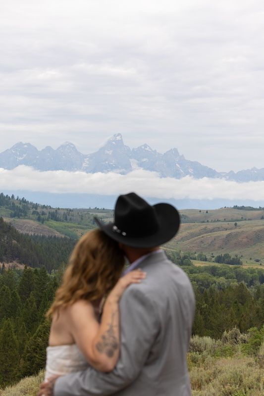 Couple in cowboy hat embraces overlooking Grand Teton mountain range with rolling hills and evergreen forest below.