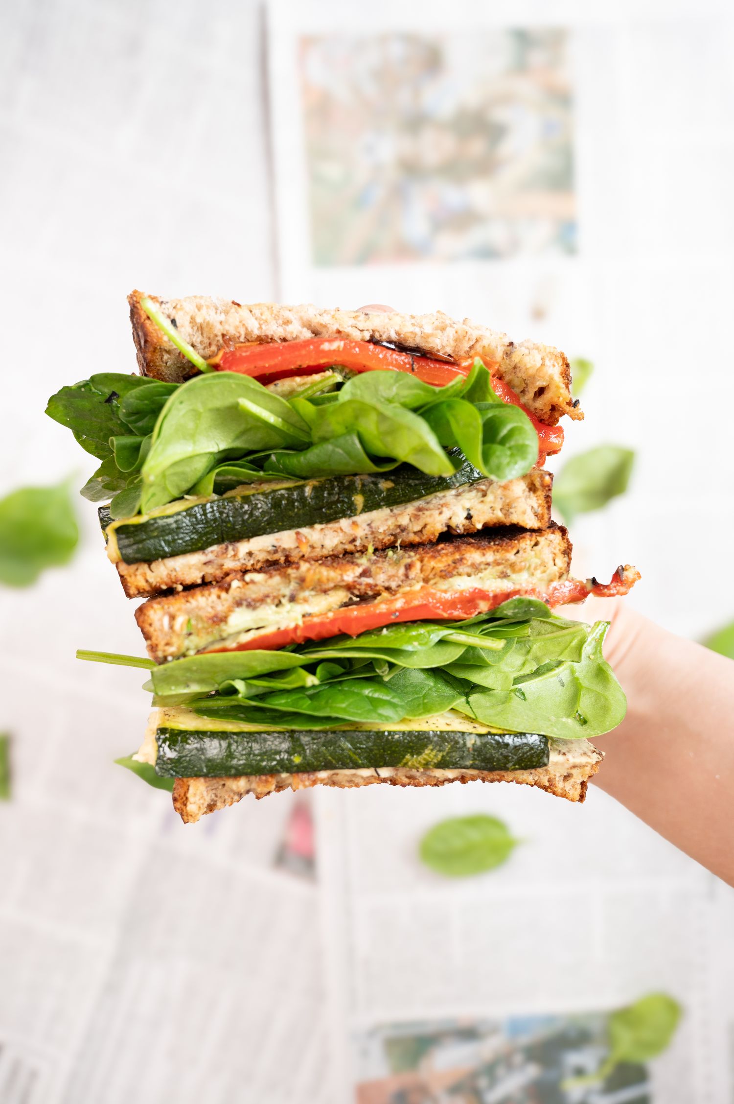 A hand holds a stacked vegetable sandwich with spinach, tomato, and zucchini on grain bread.