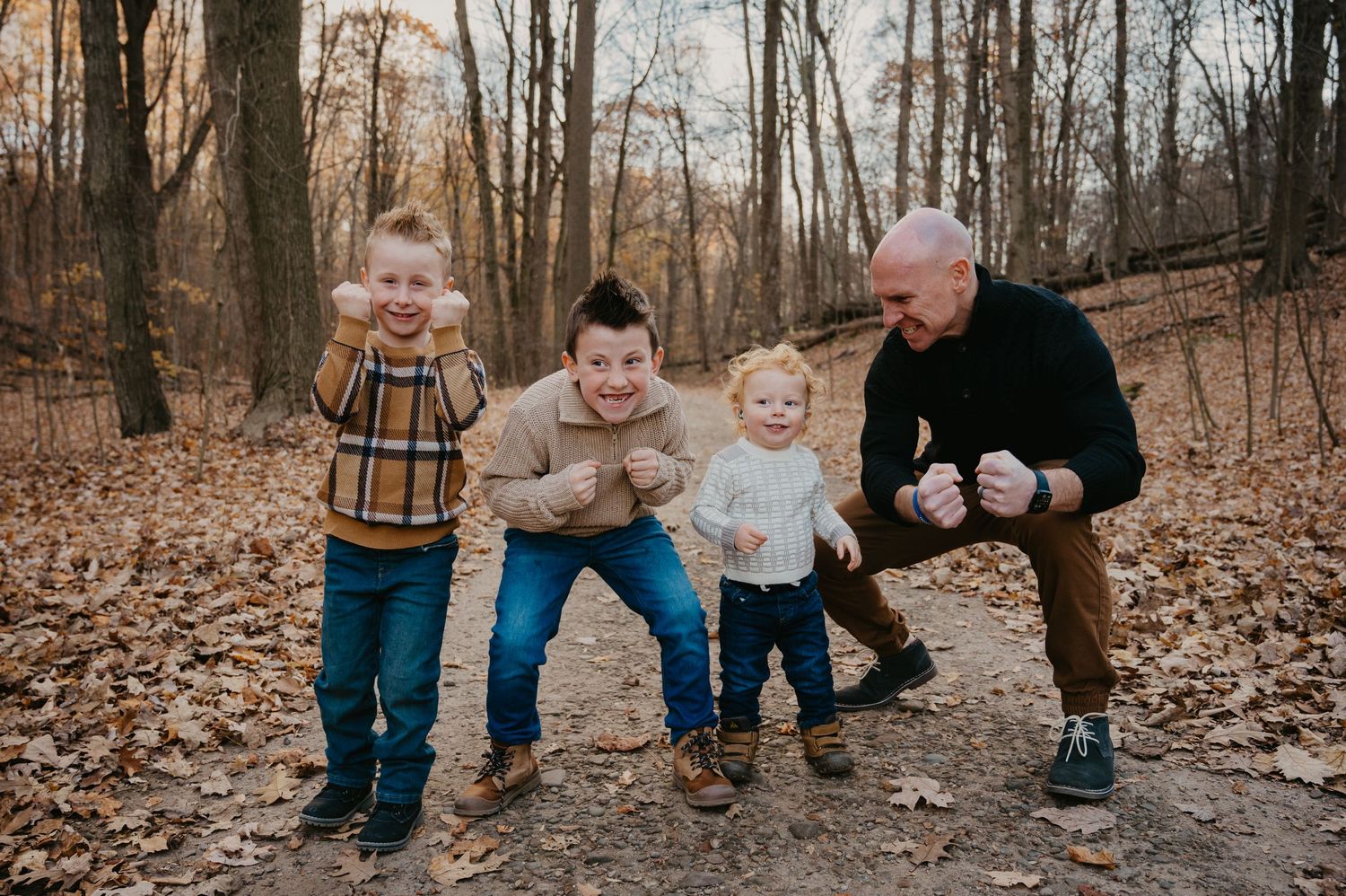 A joyful family of four strikes fun poses among autumn leaves in a wooded park setting.