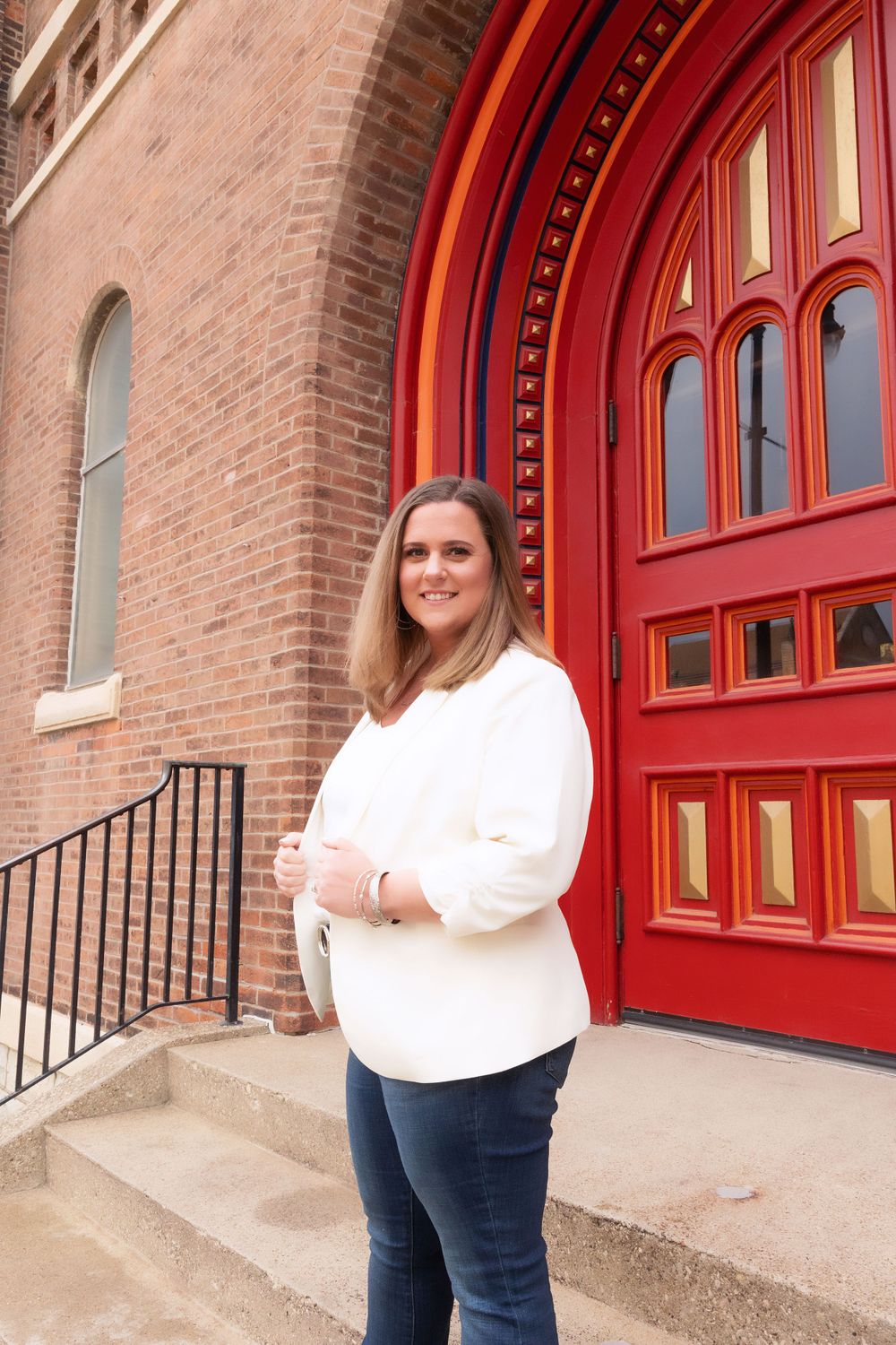Professional portrait of someone in a white blouse and jeans standing by a grand red church door.