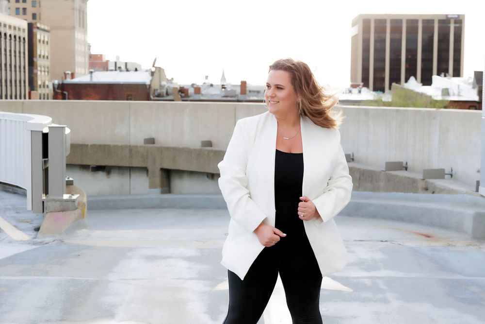 Professional woman in white blazer and black outfit poses confidently on urban rooftop with city buildings in background.