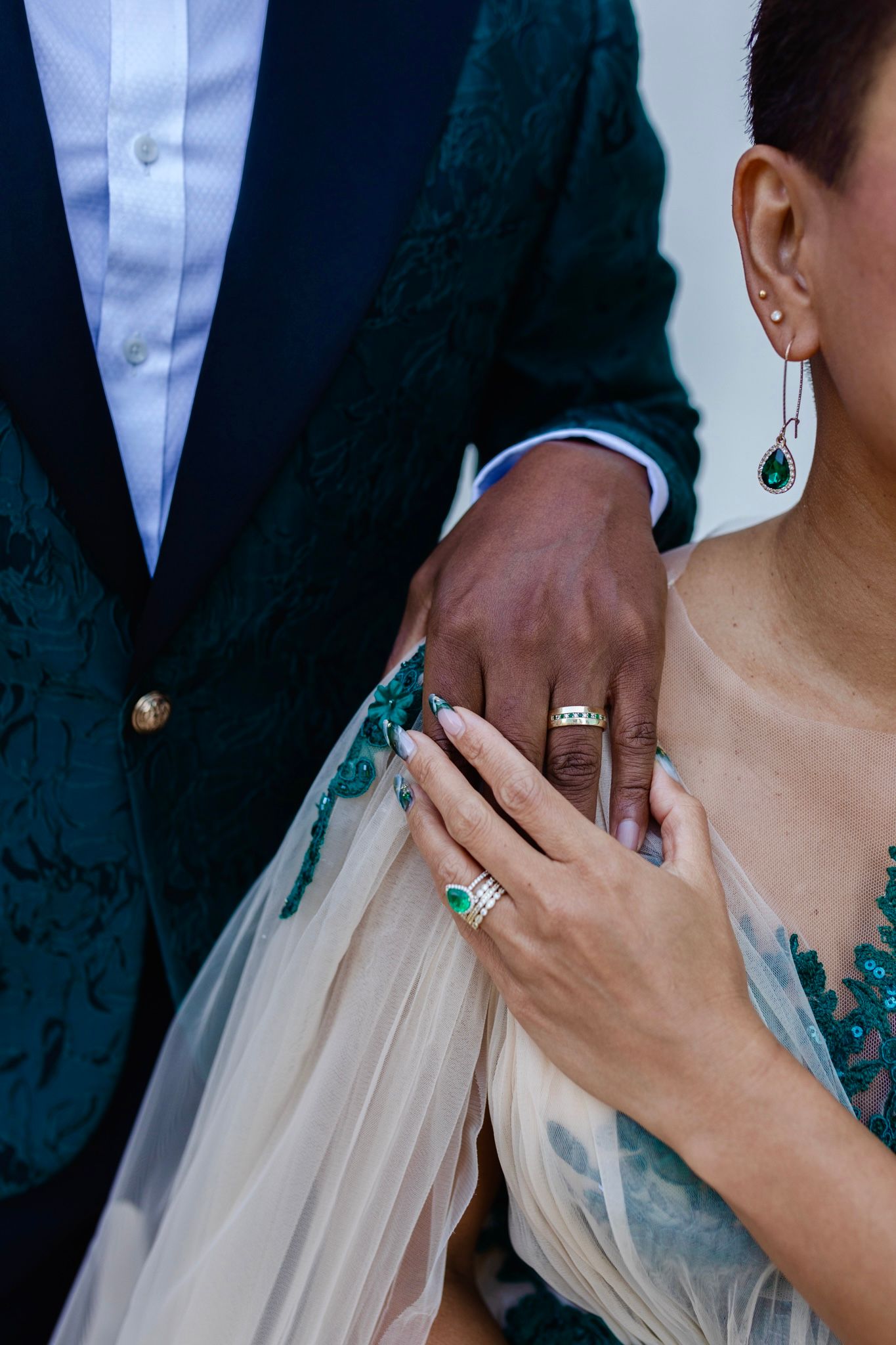 Close-up detail of wedding attire showing emerald green details, rings, and delicate white fabric.