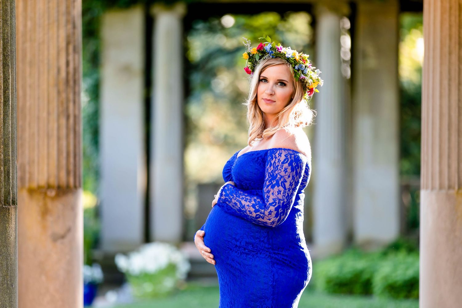 An expectant mother in a royal blue dress poses between classical columns wearing a floral crown.