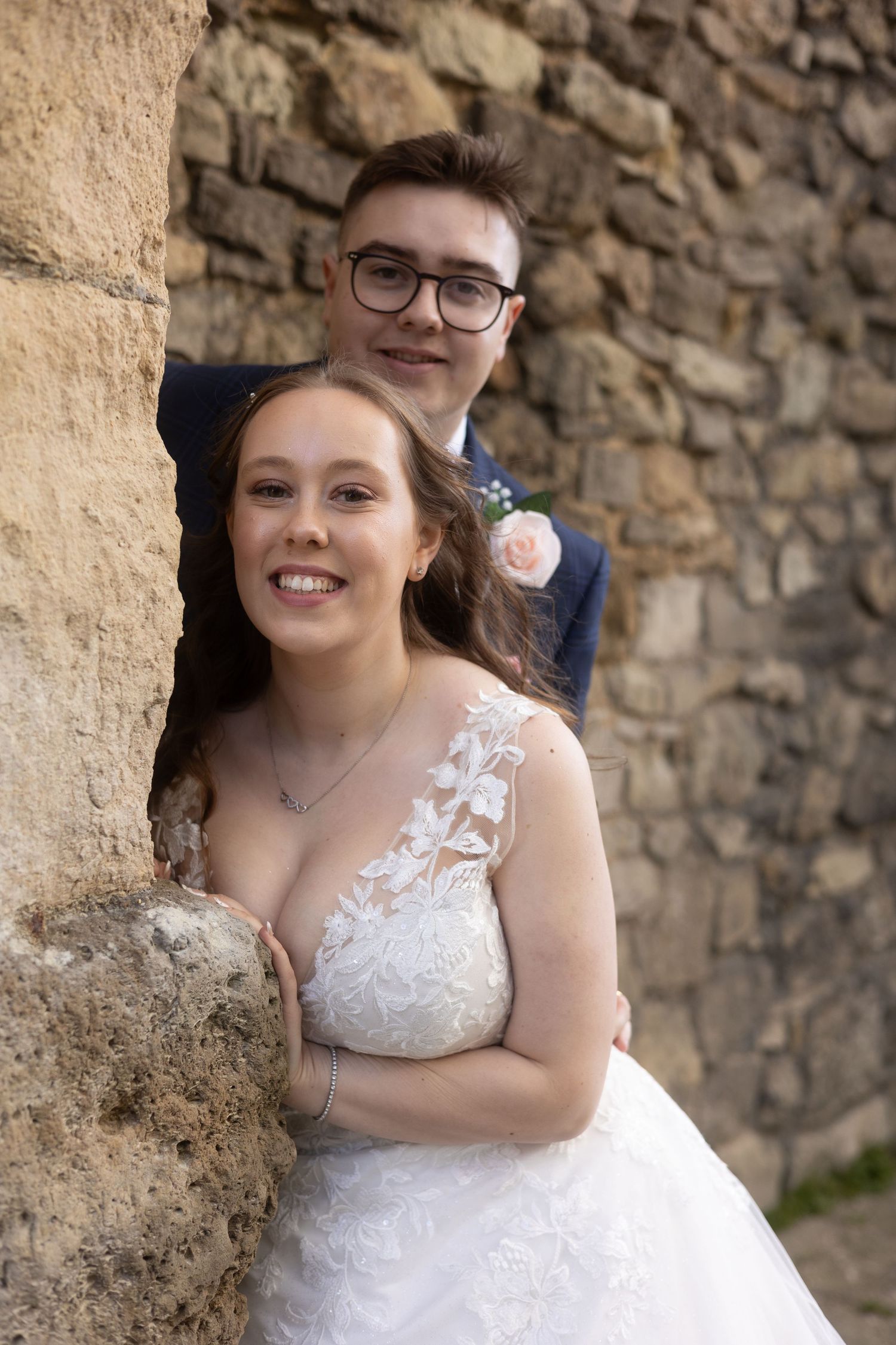 Wedding couple posing against rustic stone wall with delicate lace dress details.