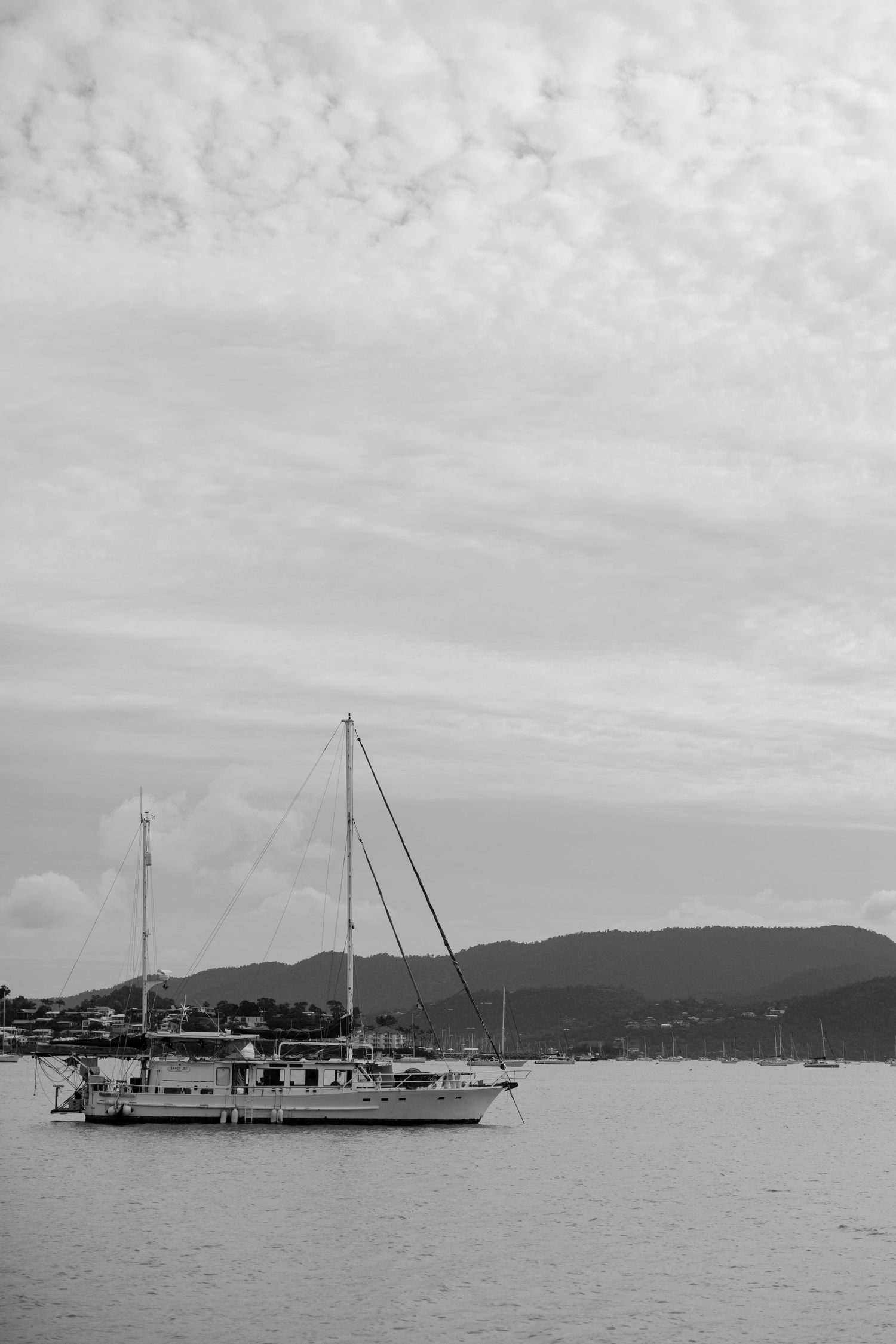 Black and white image of sailboats anchored in a harbor with mountains in background.