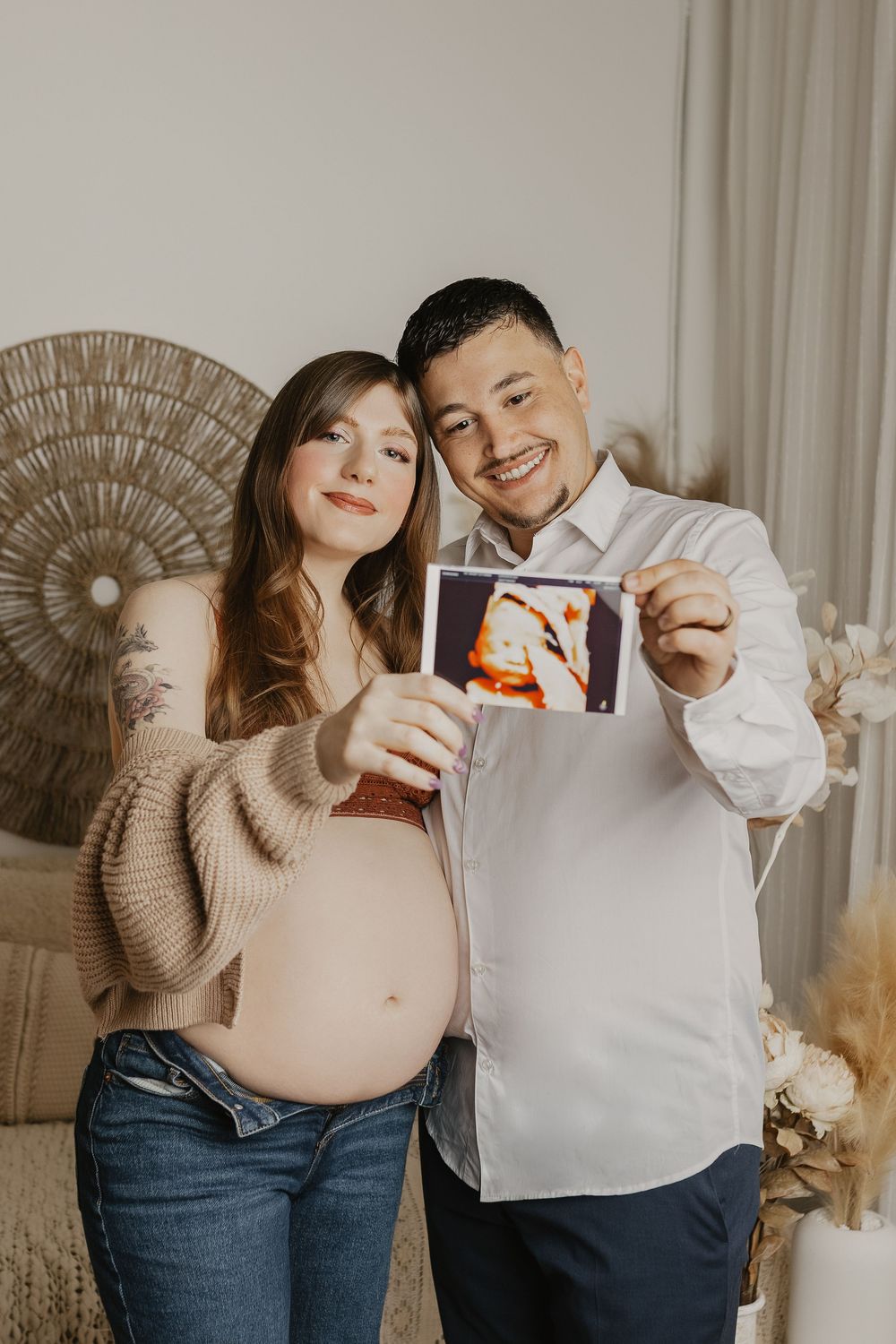 Smiling pregnant couple holding ultrasound photo together in cozy room, woman showing baby bump in jeans and cardigan.