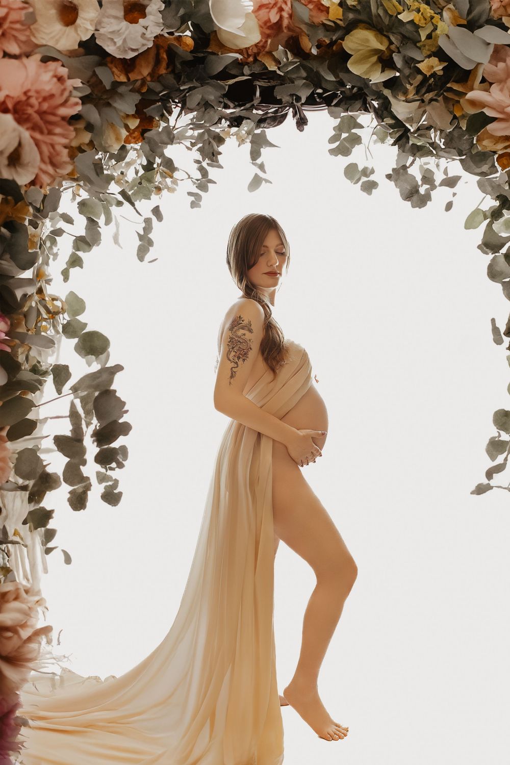 Pregnant woman draped in sheer fabric stands under floral arch with eucalyptus, showcasing tattoo in maternity photo.