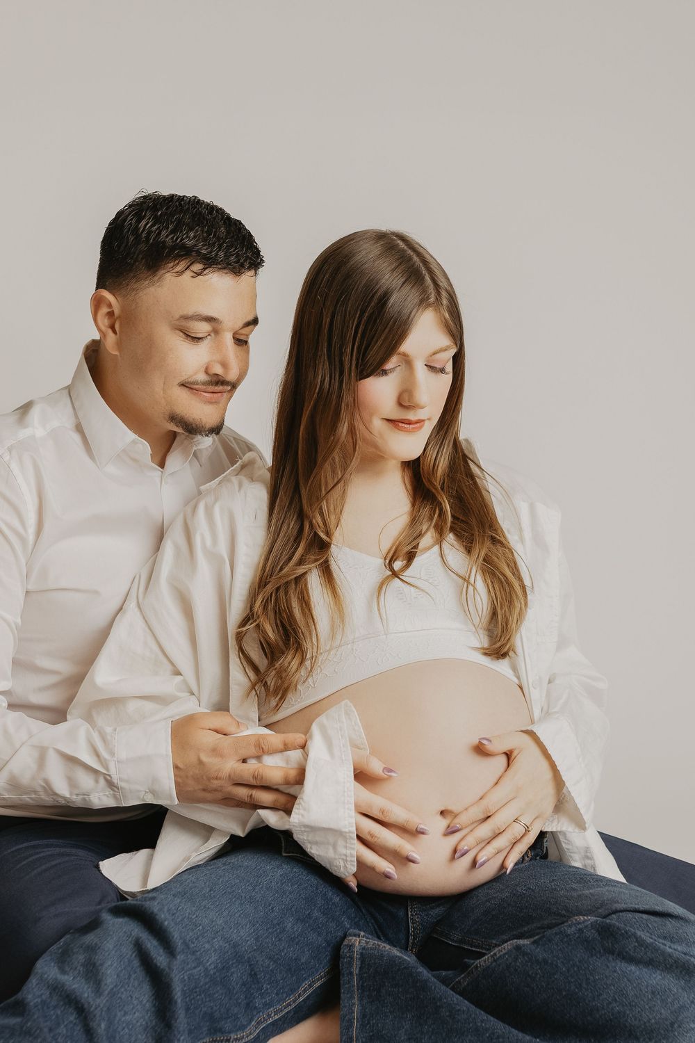 A couple tenderly cradles a pregnant belly, both dressed in white shirts and jeans against a neutral background.