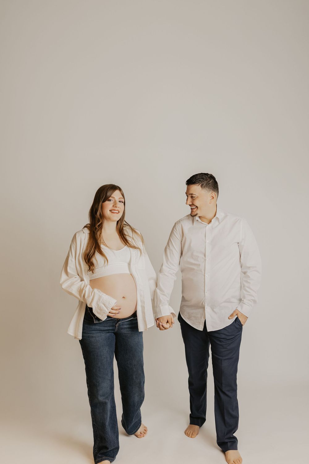 Smiling pregnant couple holding hands in a bright studio, wearing white shirts and jeans during a maternity photoshoot.