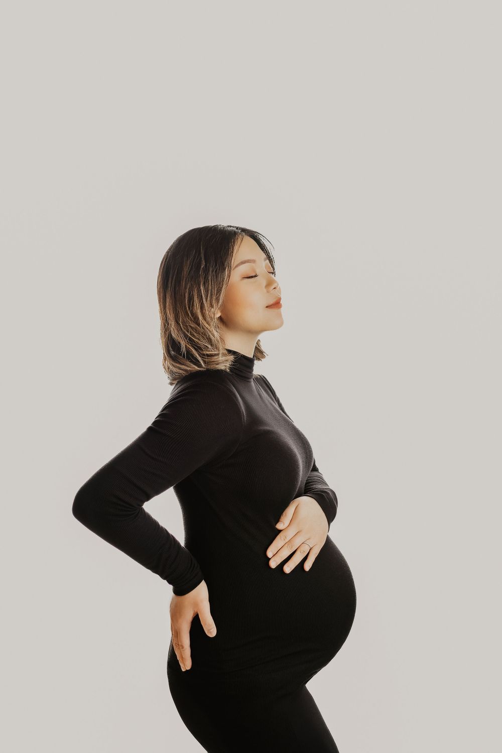Pregnant woman in black outfit cradling baby bump with eyes closed against light gray background.