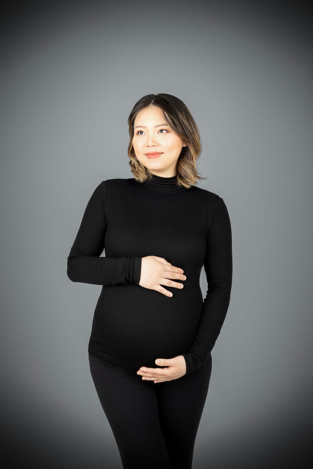 Pregnant woman in black turtleneck outfit cradling baby bump, smiling against gray studio background.