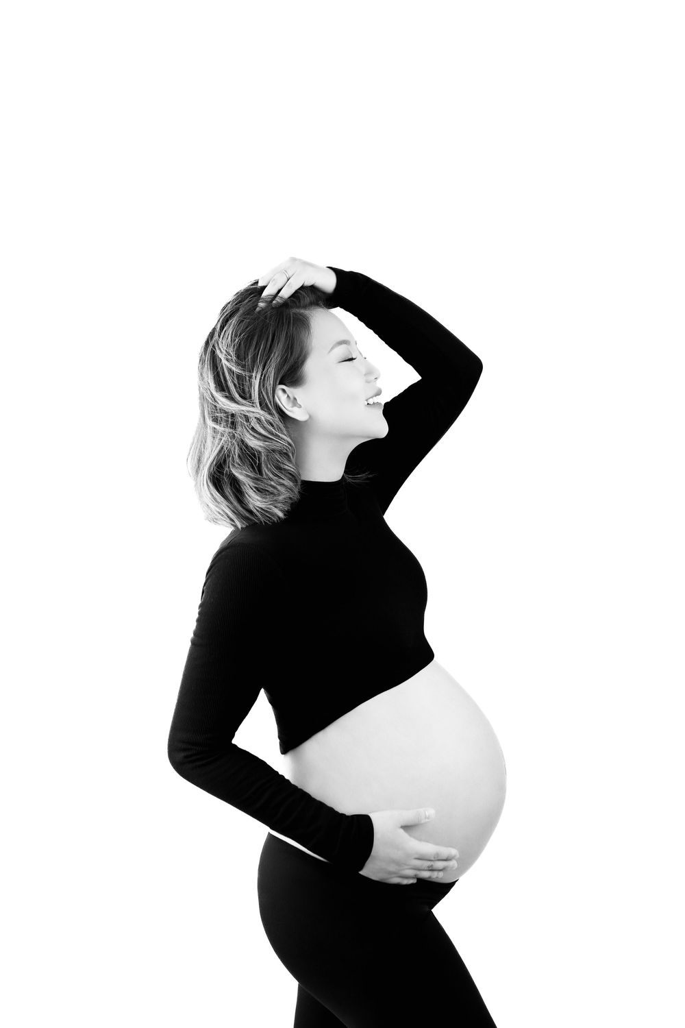 Black and white photo of a smiling pregnant woman in black outfit cradling her baby bump against white background.