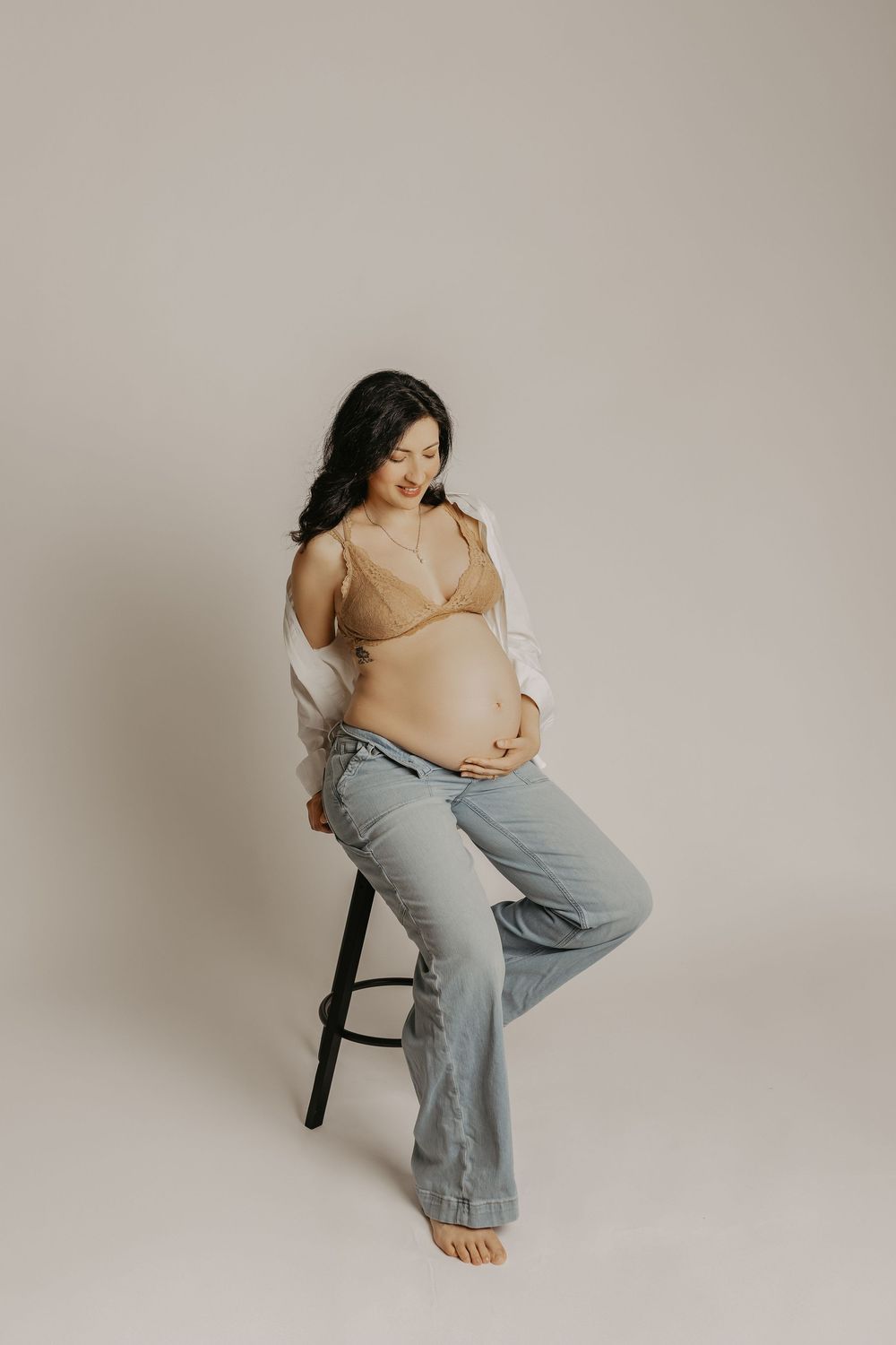 Pregnant woman in beige bralette, open white shirt, and jeans sitting on stool, cradling baby bump in studio.