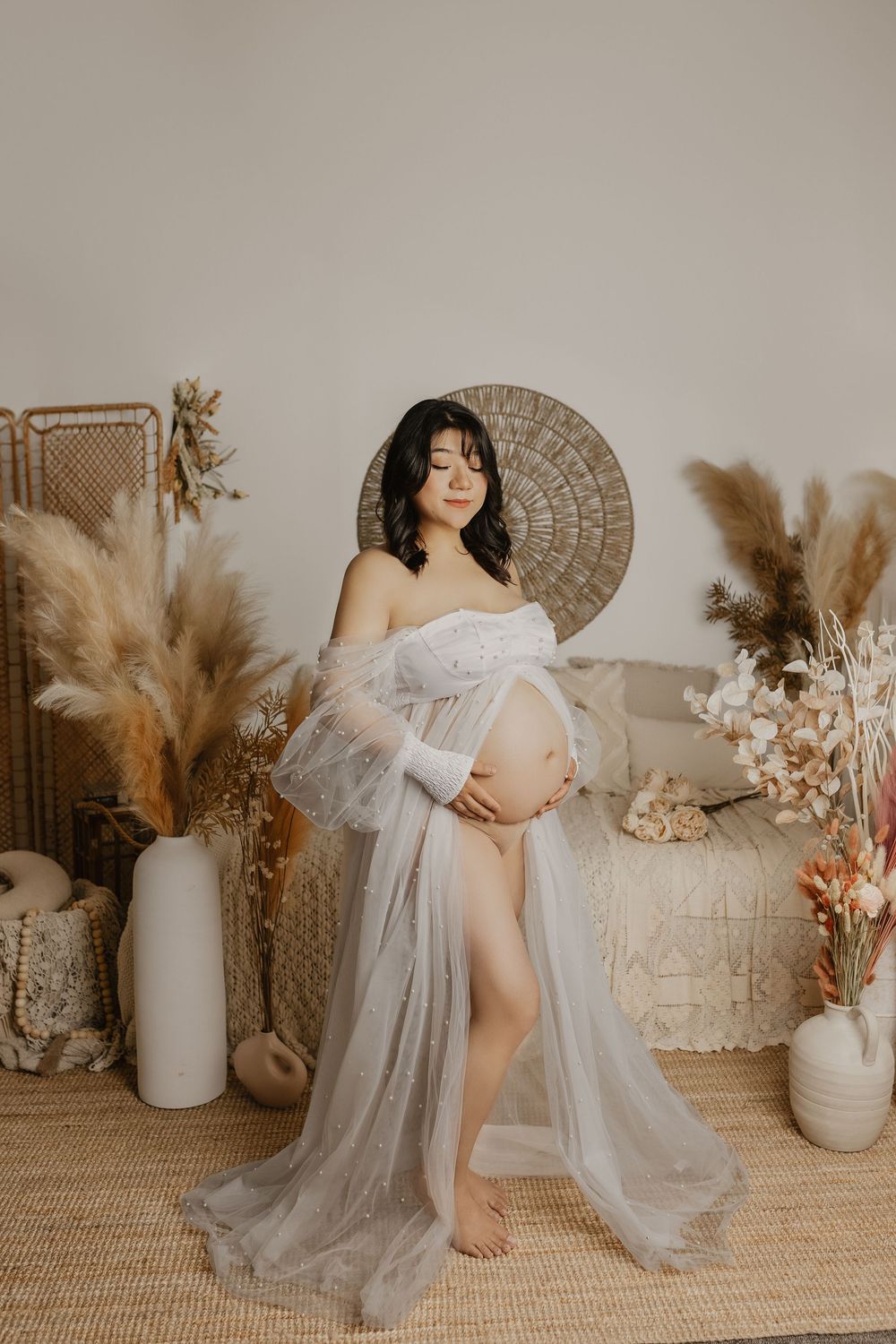 Pregnant woman in sheer white lace robe poses in boho studio with pampas grass and rattan decor.