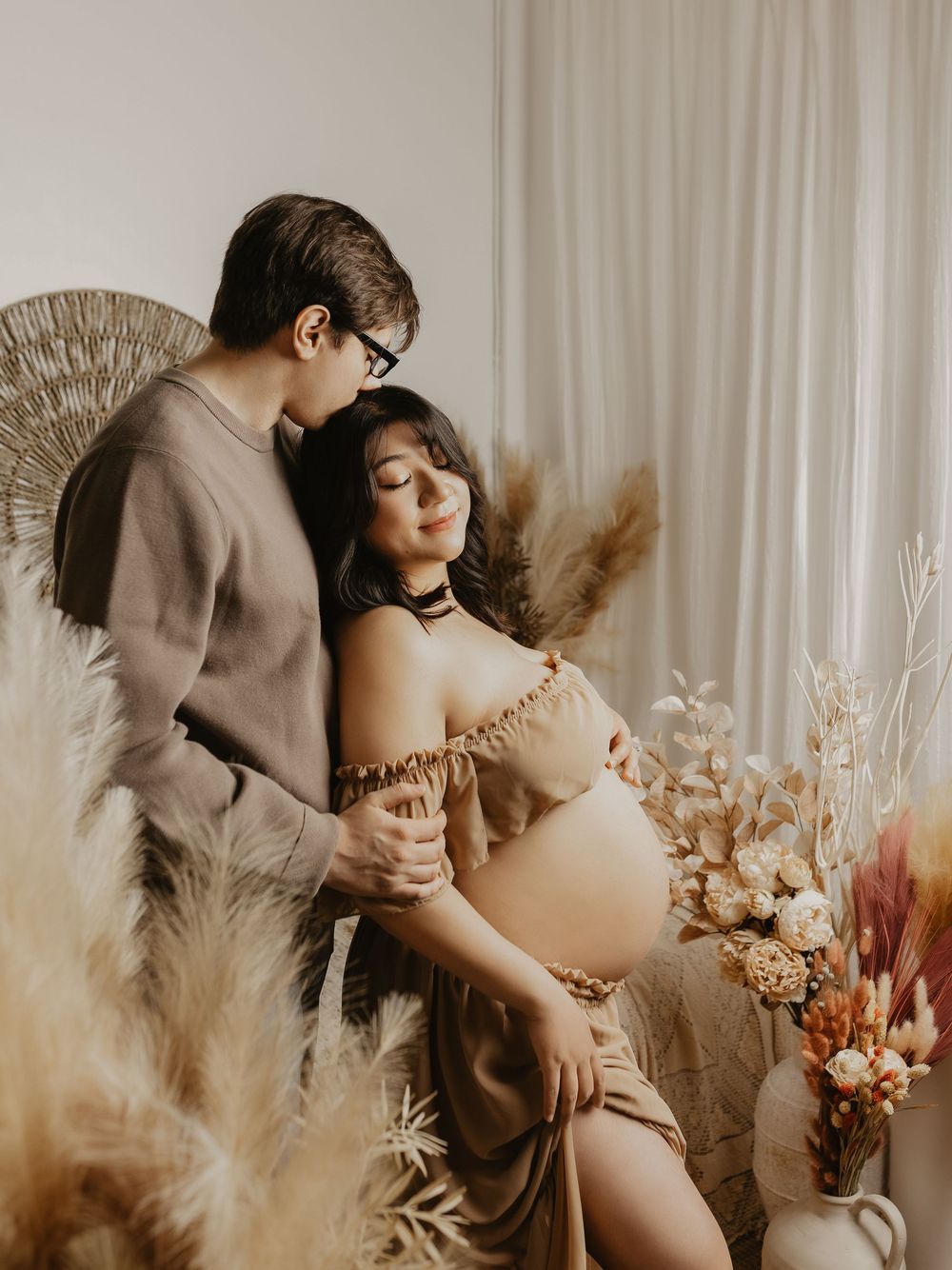 A couple shares a tender maternity photo moment surrounded by dried pampas grass and floral arrangements in warm neutral tones.