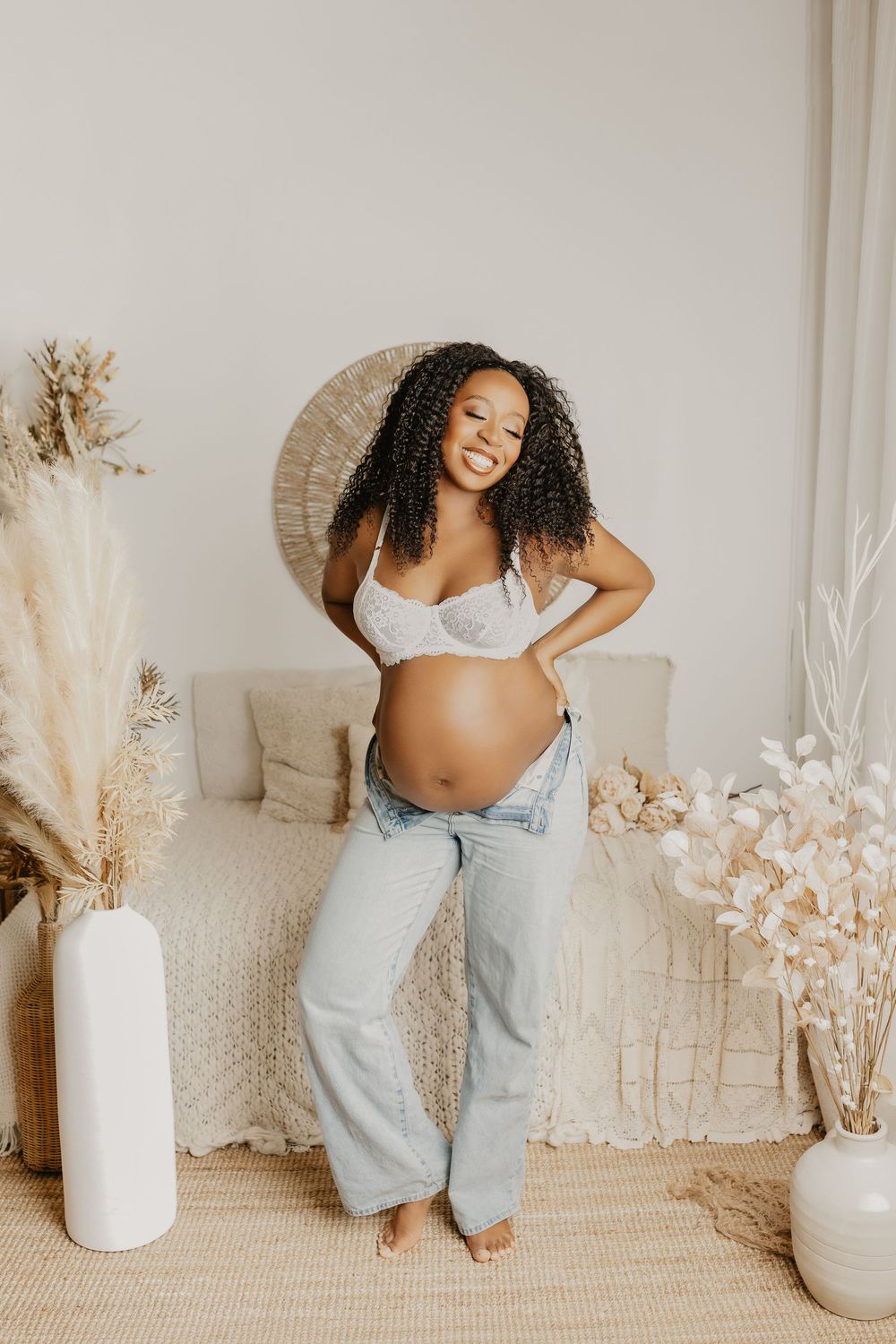 Smiling pregnant woman in lace bralette and unzipped jeans showing baby bump in boho-styled room with pampas grass.