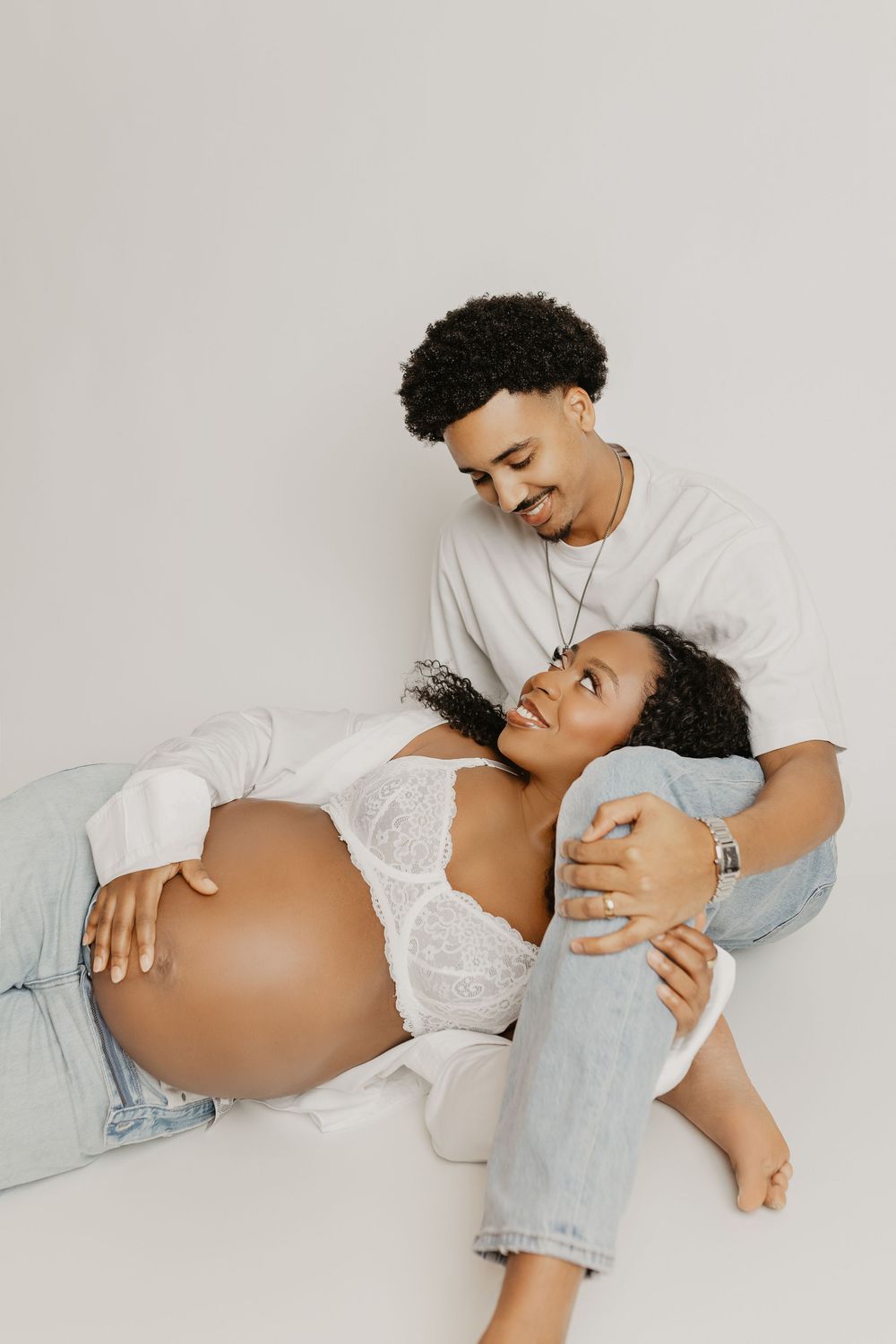 A pregnant couple shares a tender moment, with the woman lying down as her partner smiles at her lovingly in a bright studio.