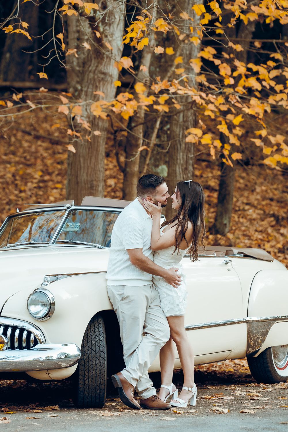 Romantic couple poses by vintage white car with autumn foliage in background. Pittsburgh Wedding Photography, Videography