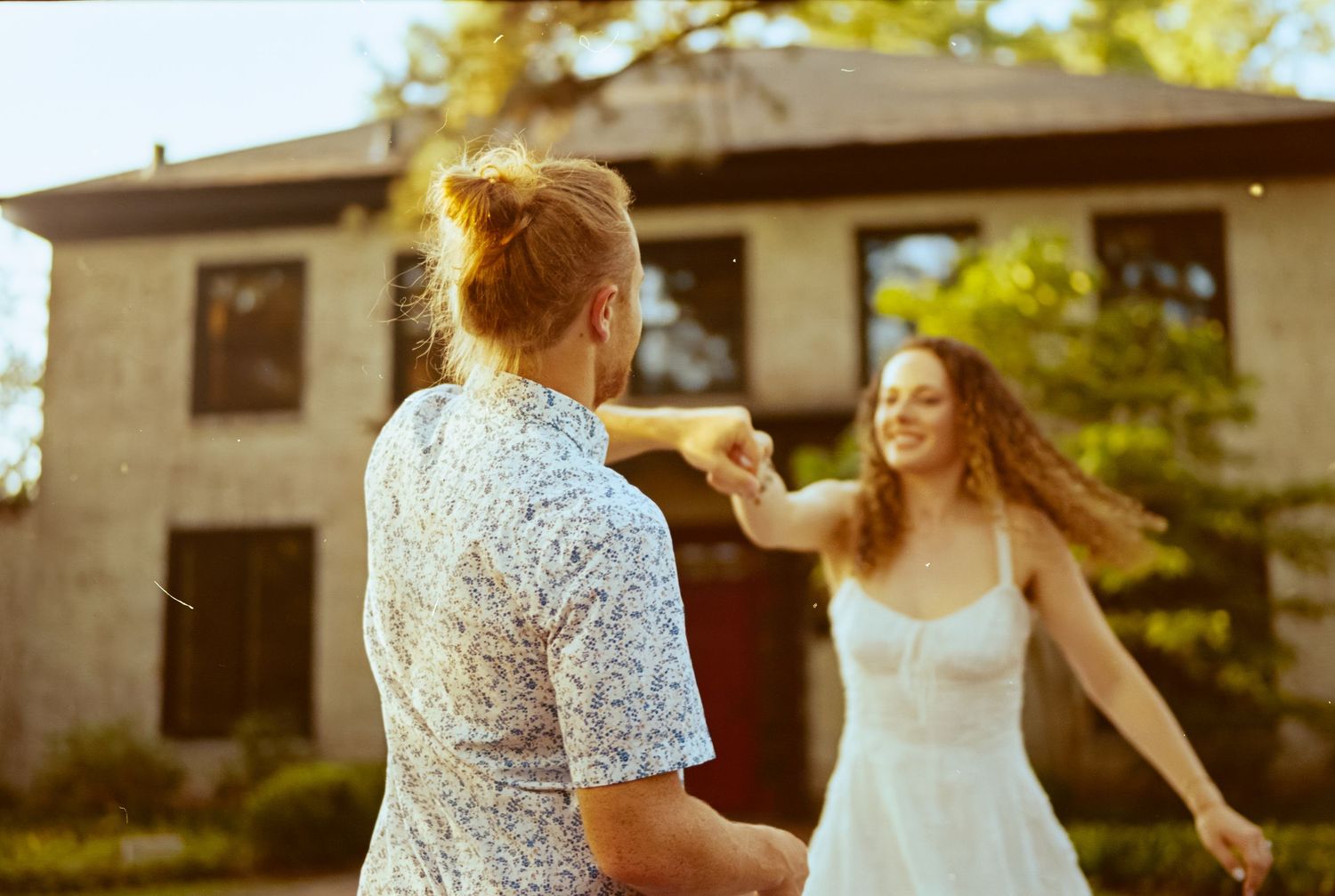 A bride and bridesmaid share a candid moment during an outdoor wedding celebration in warm sunlight.