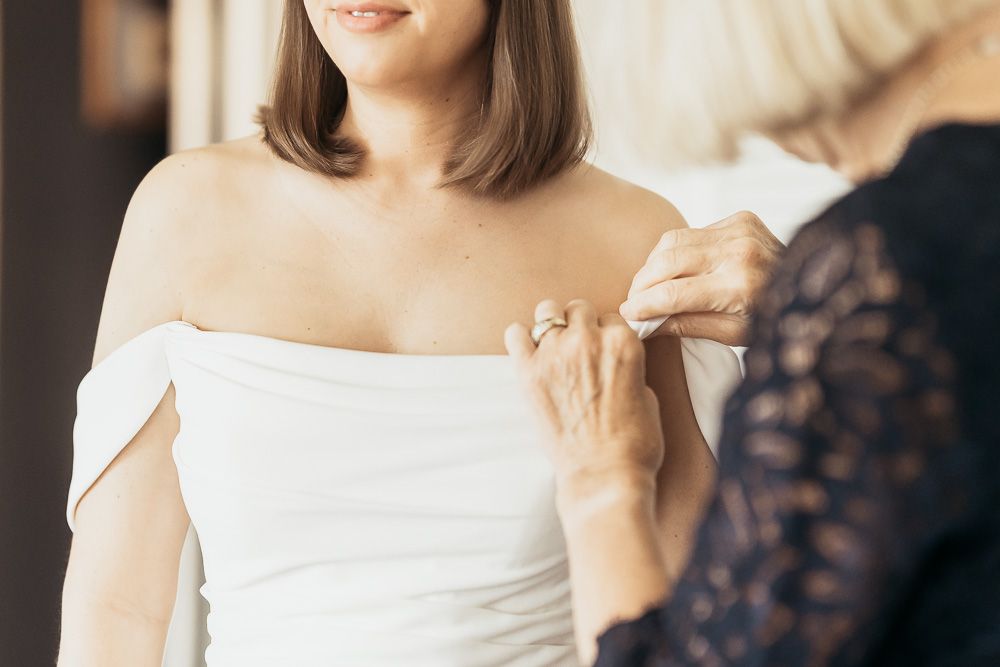 Someone in a white off-shoulder wedding dress being zipped up by mom for wedding photography and videography