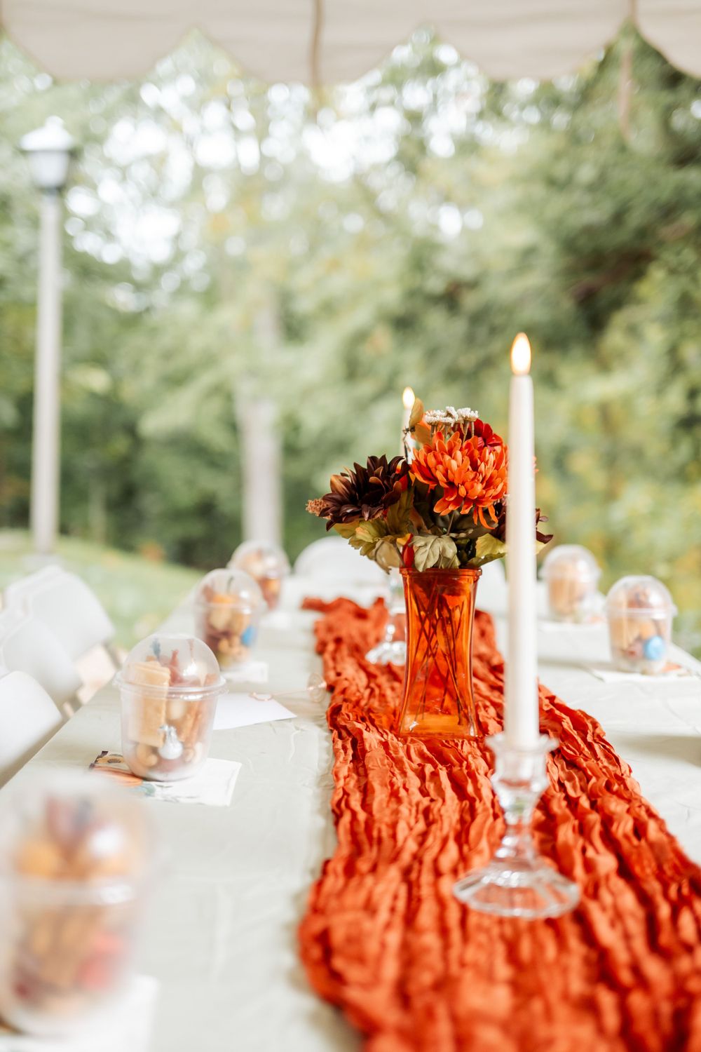 An elegant table setting with burnt orange fabric runner, white candles, and autumn floral centerpieces at an outdoor event.
