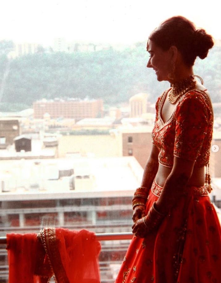 A silhouette stands pensively by a window in a red traditional sari, overlooking a city skyline in soft light. Hindi Cultural Wedding in pit