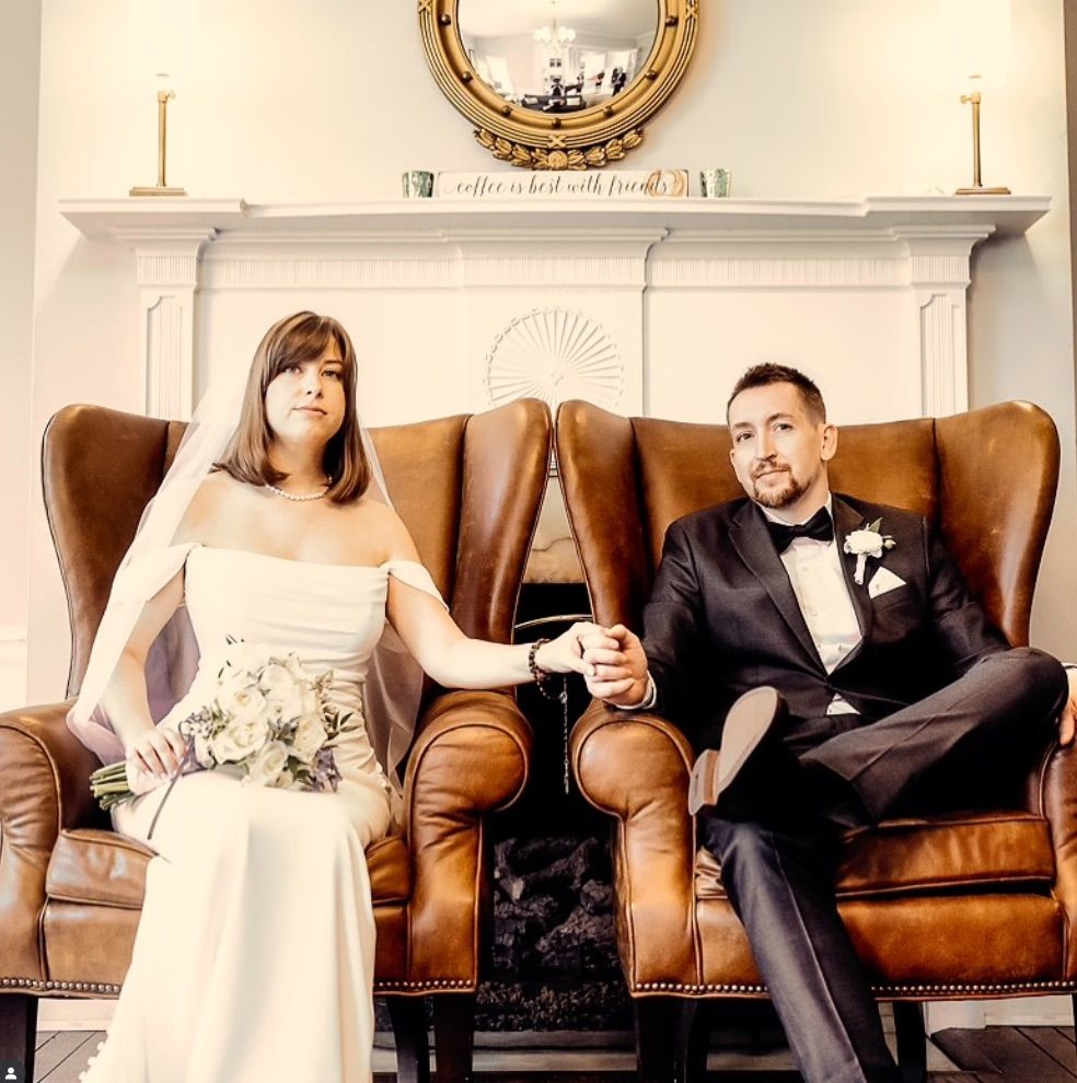 A couple in wedding attire sits on vintage brown leather armchairs in front of a white fireplace with round mirror above.