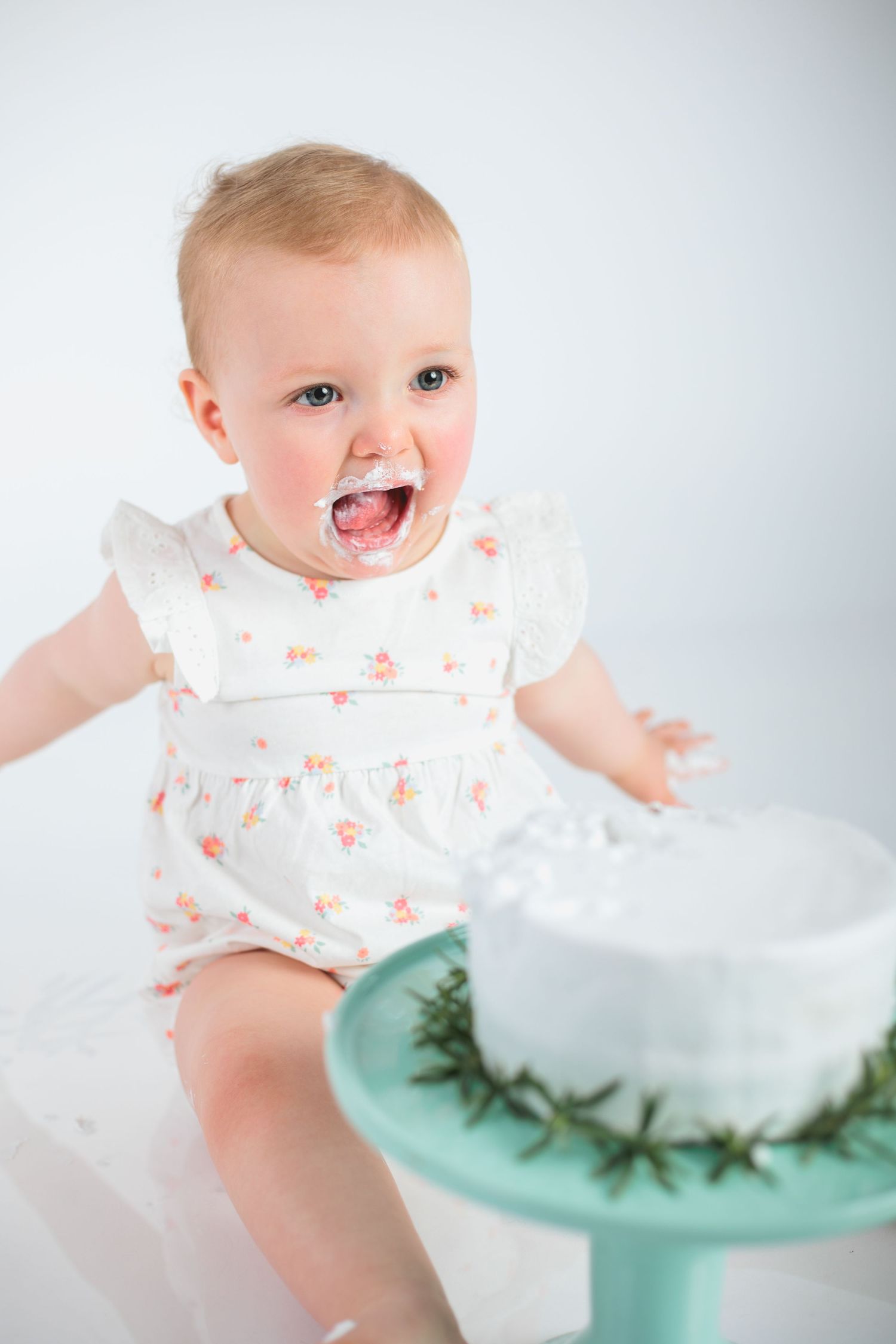 Joyful baby wearing floral white dress sits next to turquoise stand with draped fabric and greenery.