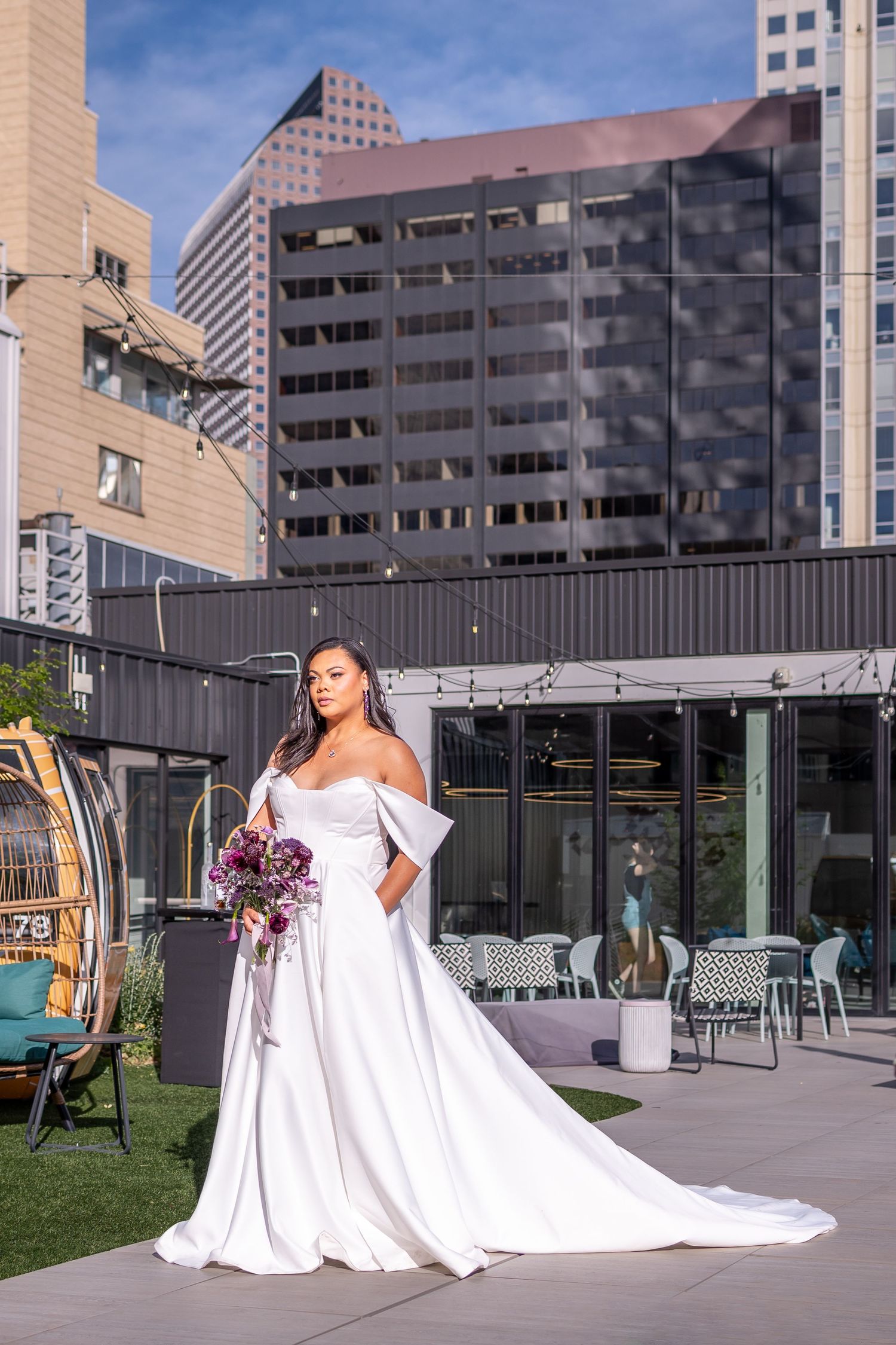 A bride in a flowing white off-shoulder gown with a long train poses with a purple bouquet on a rooftop terrace downtown Denver,Colorado