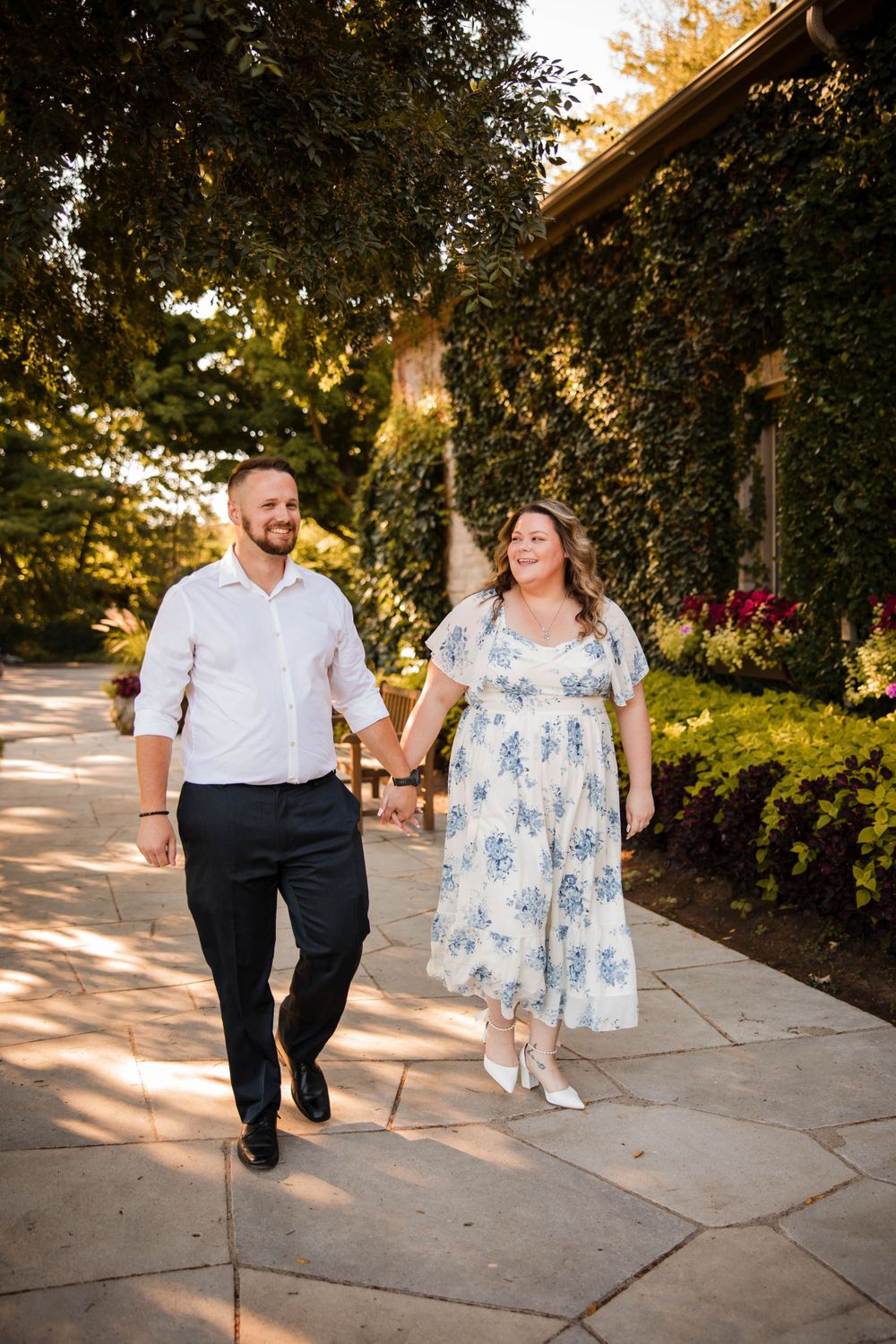 A couple walking hand in hand along a stone path surrounded by lush greenery.