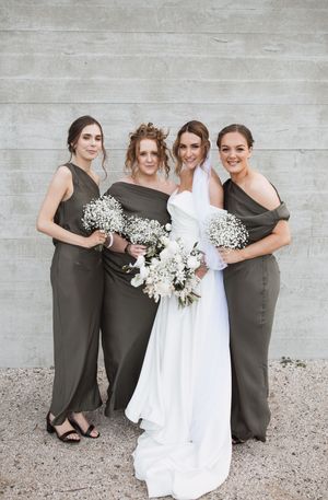 Bridal party poses together wearing white and olive green dresses against concrete wall.