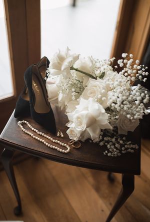 Wedding accessories including white roses, pearls, and black heels arranged on dark wooden table.