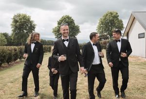 Groomsmen in black tuxedos laughing together on a cloudy day.