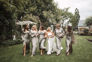 Wedding party members in grey and white attire run playfully across a green lawn.