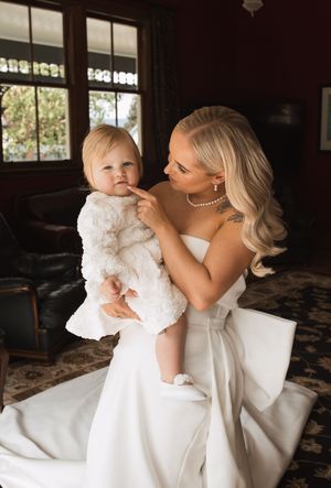 Tender moment between bride in white gown and small child in white dress indoors.