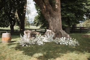 Rustic outdoor wedding ceremony setup with wooden barrels and white floral arrangements under large trees.