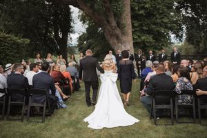 Outdoor wedding ceremony with guests seated on white chairs beneath towering trees.