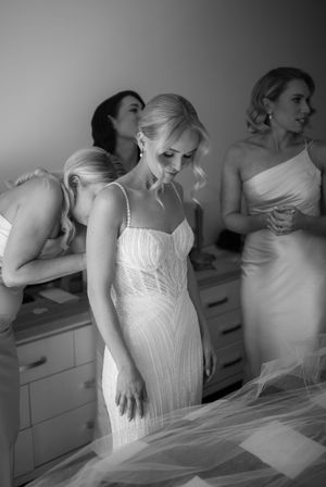 Women in white dresses prepare for a wedding in a brightly lit dressing room.