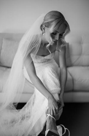 A bride in a white dress and veil adjusts her shoes in black and white photography.