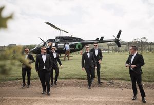 Groomsmen in black suits pose near a helicopter on a grassy field.