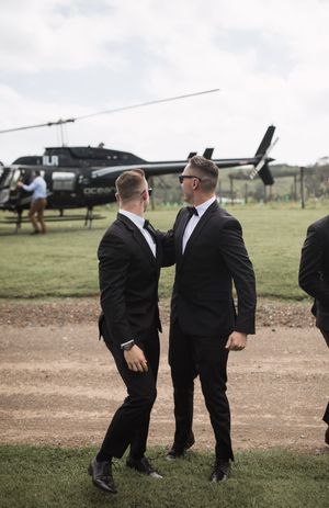 Wedding party members in black suits chat casually by a parked helicopter.