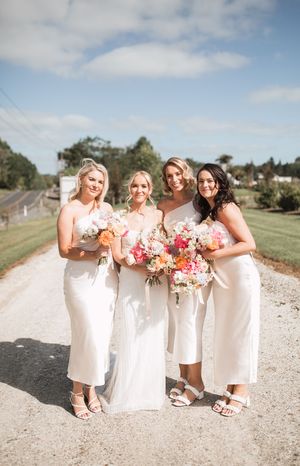 Bridesmaids in white dresses pose together holding colorful bouquets on a sunny day.